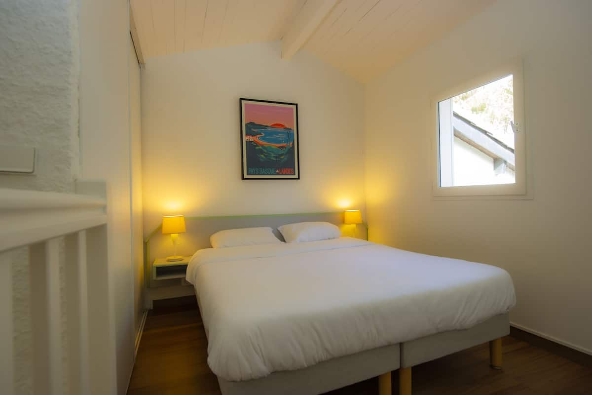 A bedroom with a bed, two bedside lamps, a painting on the wall, and a window.