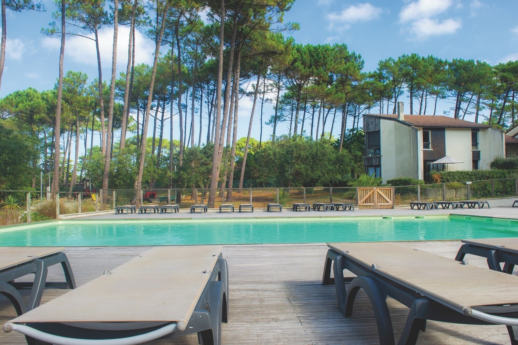 A swimming pool with lounge chairs, surrounded by tall trees and a building with a tiled roof.