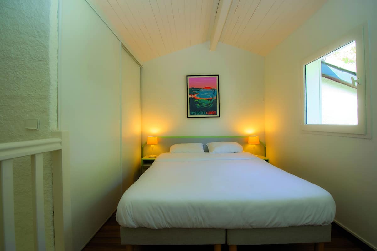 A neatly made bed in a room with a single painting on the wall, two bedside lamps, and a window on the right.