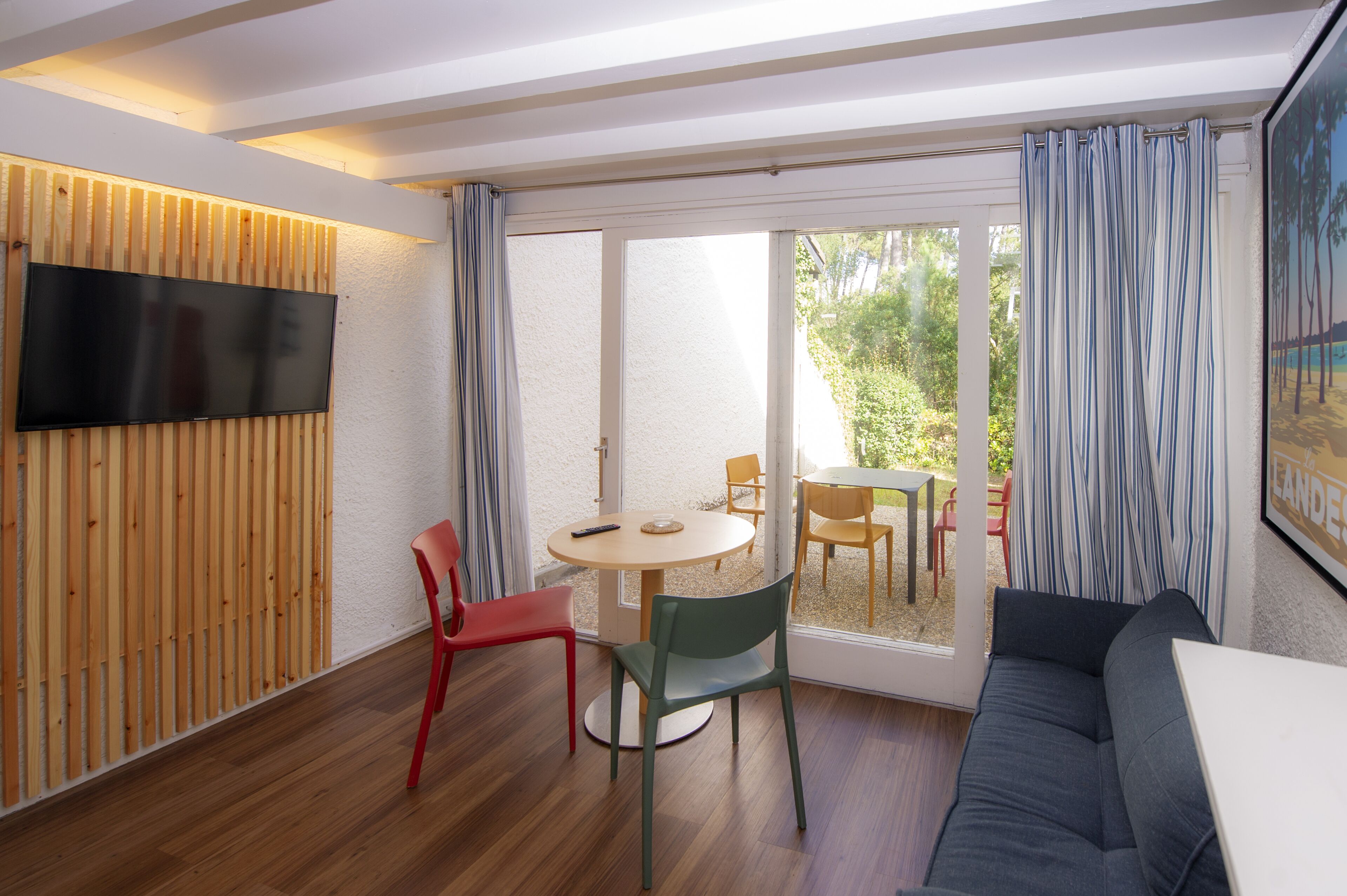 Een moderne kamer met een televisie bevestigd aan een houten paneelwand, een ronde tafel met stoelen, een bank en een schuifdeur van glas naar een buitenruimte.