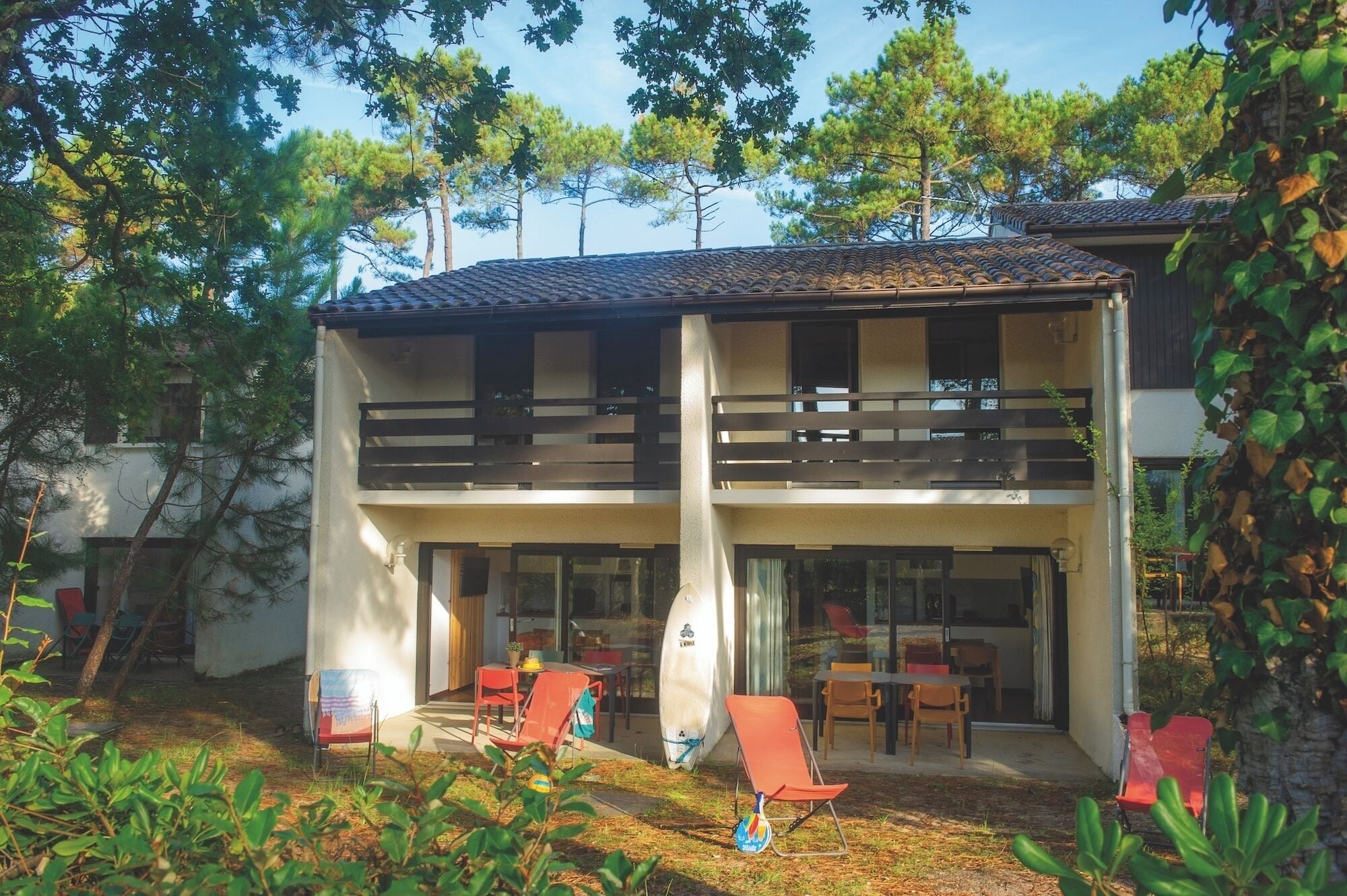Un edificio de dos pisos con balcón, asientos al aire libre y una tabla de surf.