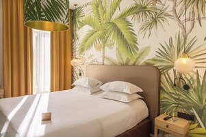 Un lit bien fait, avec du linge de lit blanc, une table de chevet en bois sur laquelle repose une télécommande, et un mur orné d’une fresque représentant des plantes tropicales.