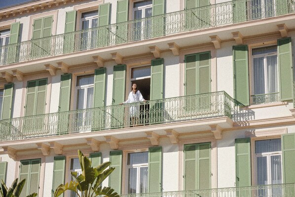 Ein mehrstöckiges Gebäude mit Balkonen und grünen Fensterläden, auf einem Balkon steht eine Person.