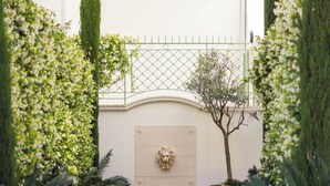 A narrow pathway flanked by tall, manicured hedges leading to a building with a decorative door and a stone fountain. - Hotel Verlaine (Cannes)