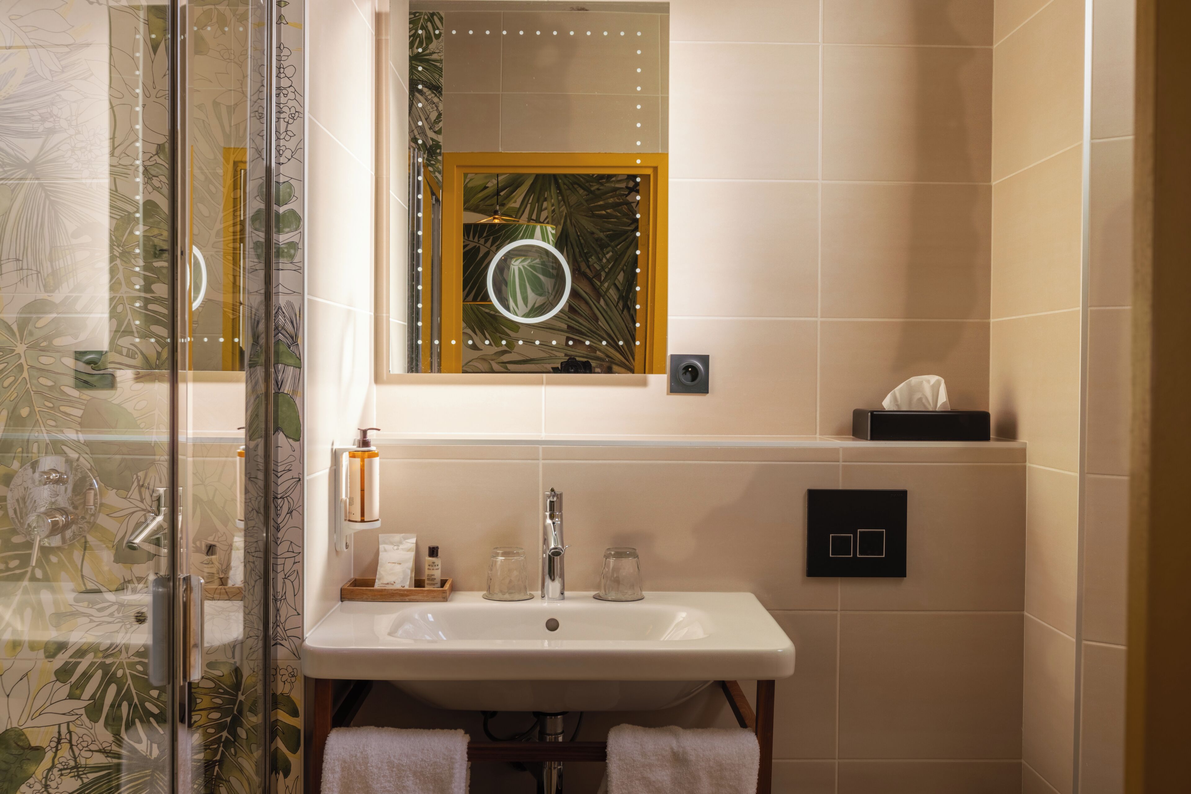 Un baño con cabina de ducha de vidrio, un lavamanos con dos frascos de vidrio, un espejo con marco circular e interruptor de luz en la pared.