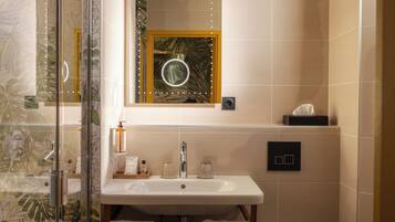 Un baño con cabina de ducha de vidrio, un lavamanos con dos frascos de vidrio, un espejo con marco circular e interruptor de luz en la pared.