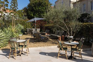 Un patio al aire libre con mesas y sillas redondas, rodeado de follaje y un edificio al fondo.