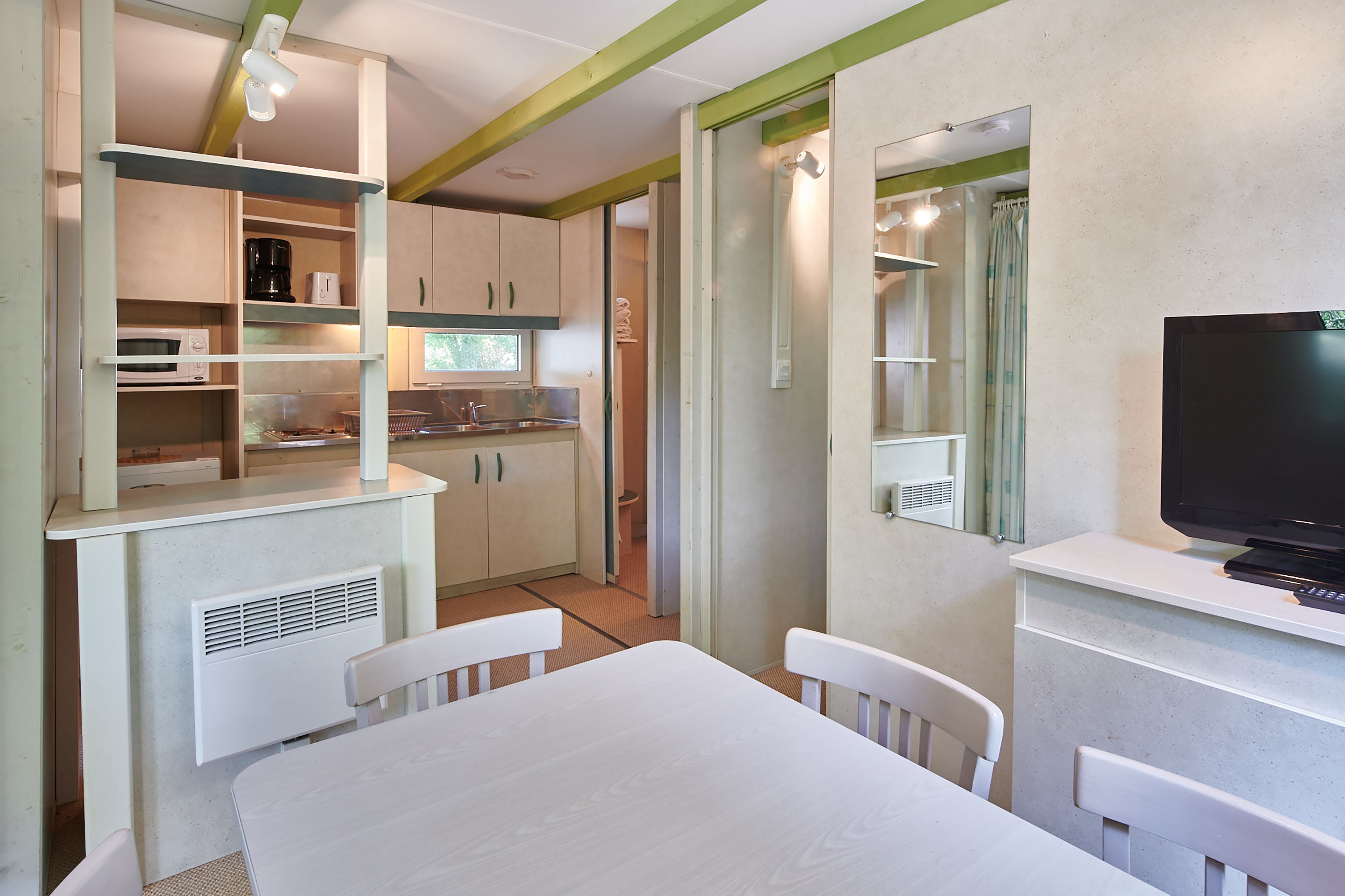 Een kleine keuken met witte kasten, een magnetron en een spoelbak. Er staat een eettafel met stoelen en een televisie op een dressoir.