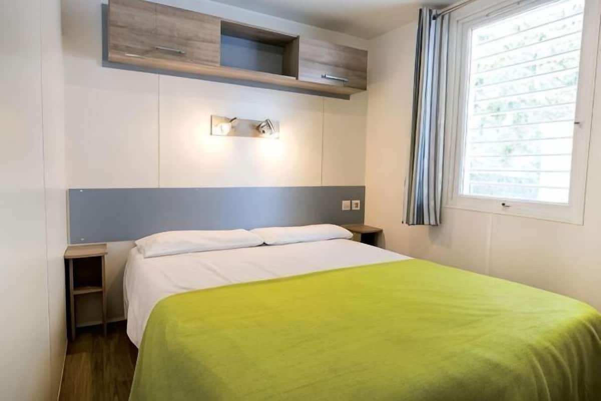 A hotel room with a bed, a nightstand, a built-in shelf above the bed, a window with curtains, and a wall-mounted light fixture.
