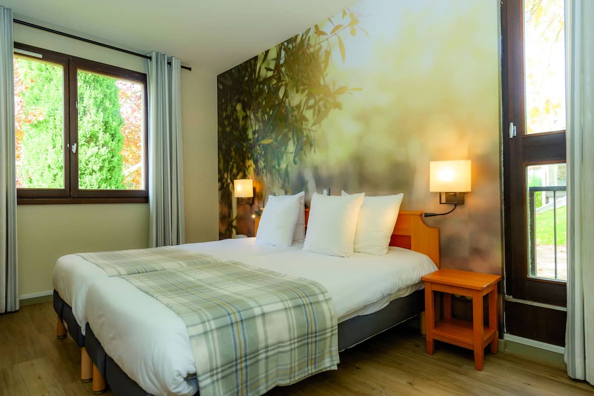 A bedroom with a large bed, two bedside lamps, a small wooden nightstand, and a window with curtains.