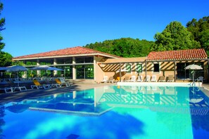 A swimming pool with lounge chairs and umbrellas, surrounded by a building with a tiled roof and glass windows.
