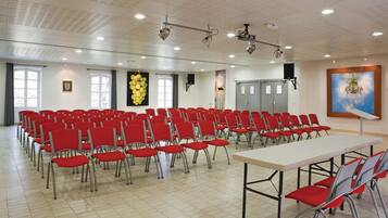 Un salón de conferencias con filas de sillas rojas, una mesa blanca y proyectores en el techo.