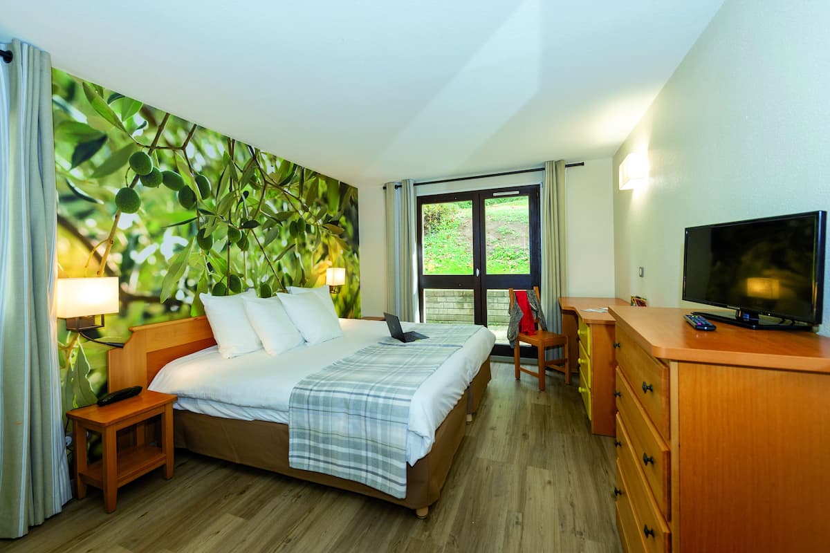 A hotel room with a bed, a desk, a television, and a view of greenery through the window.