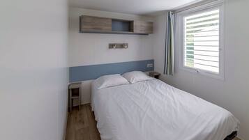 Ein Schlafzimmer mit einem Bett, einem Nachttisch, einem Holzregal über dem Bett, einem Fenster mit Jalousien und einem Lichtschalter an der Wand.
