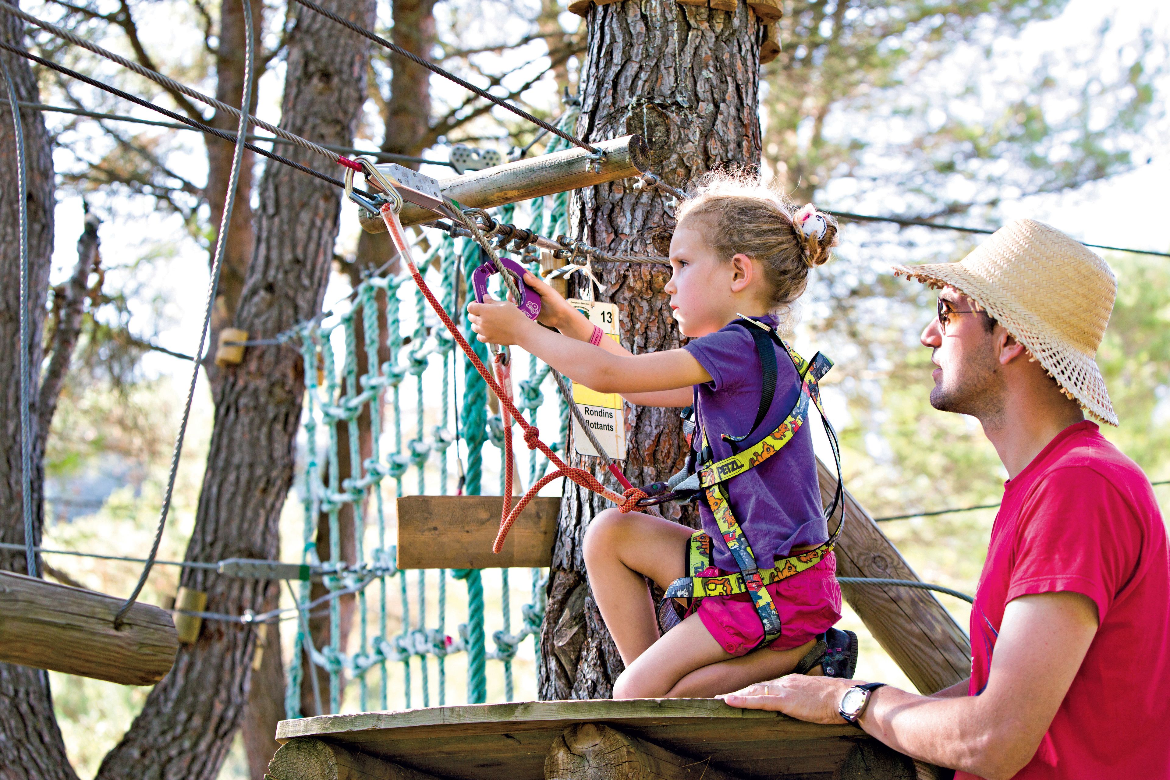 A person in a red shirt and straw hat sitting on a wooden platform, assisting a child in a purple shirt and pink shorts on a rope bridge.