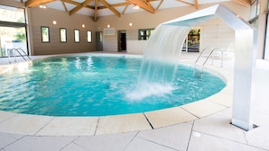 A large indoor swimming pool with a waterfall feature, wooden beams on the ceiling, and a tiled floor. - Club Vacances Bleues Domaine de Château Laval (Gréoux-les-Bains)