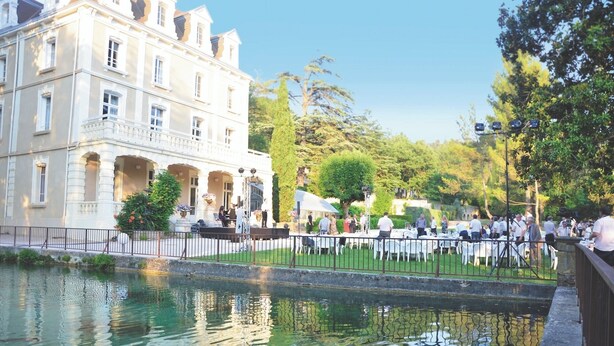 A riverside outdoor dining area with tables and chairs, a large building with classical architecture, and a calm water body reflecting the surroundings. - Club Vacances Bleues Domaine de Château Laval (Gréoux-les-Bains)