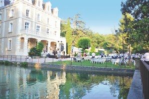 Ein Essbereich im Freien am Flussufer mit Tischen und Stühlen, ein großes Gebäude mit klassischer Architektur und ein ruhiges Gewässer, das die Umgebung spiegelt.