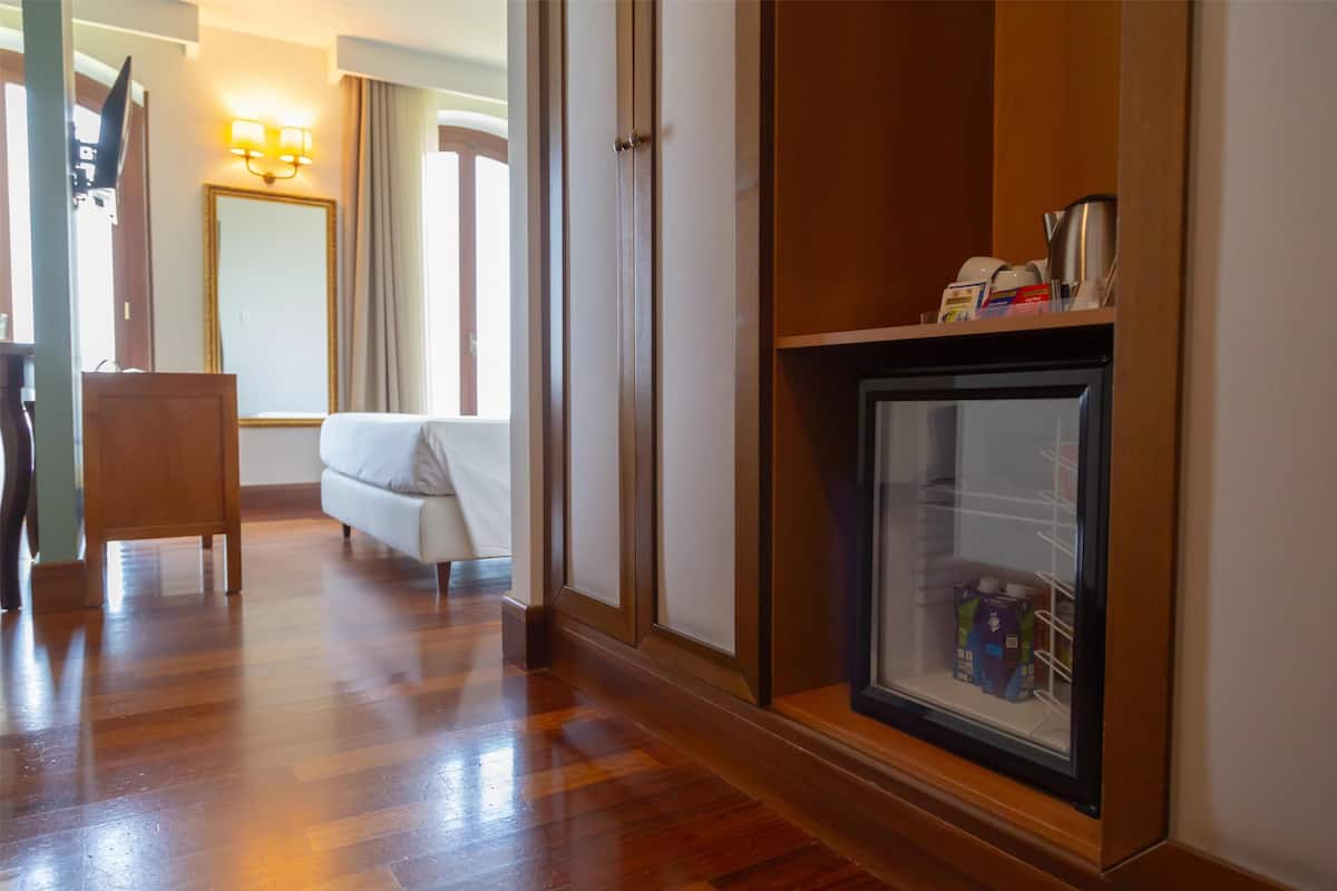 Une chambre d’hôtel avec un plancher en bois, un canapé blanc, un meuble en bois doté d’une vitrine, et une porte donnant sur une autre pièce.
