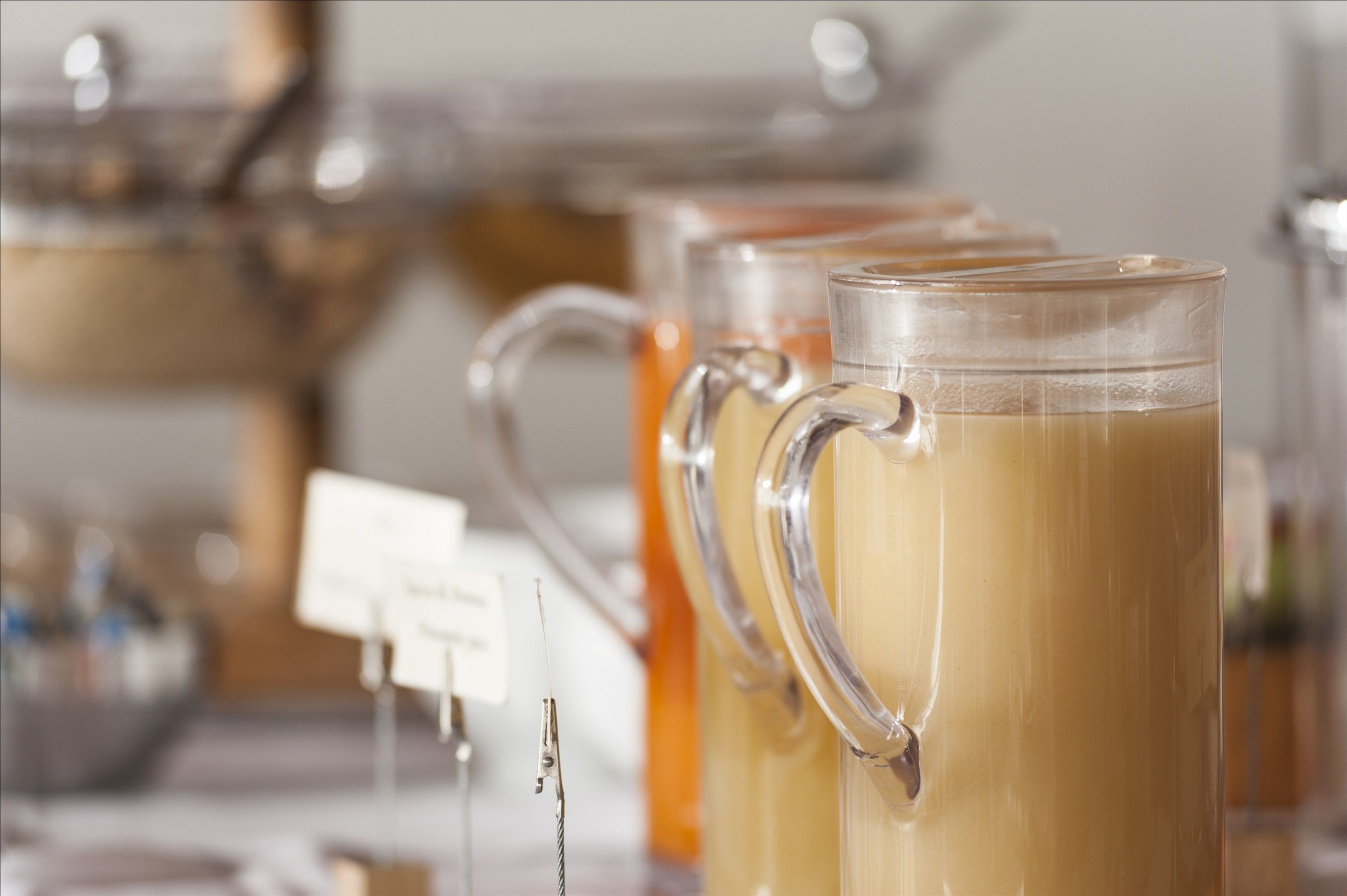 A row of clear glass mugs filled with a light-colored beverage, likely tea or coffee, on a table with a blurred background.