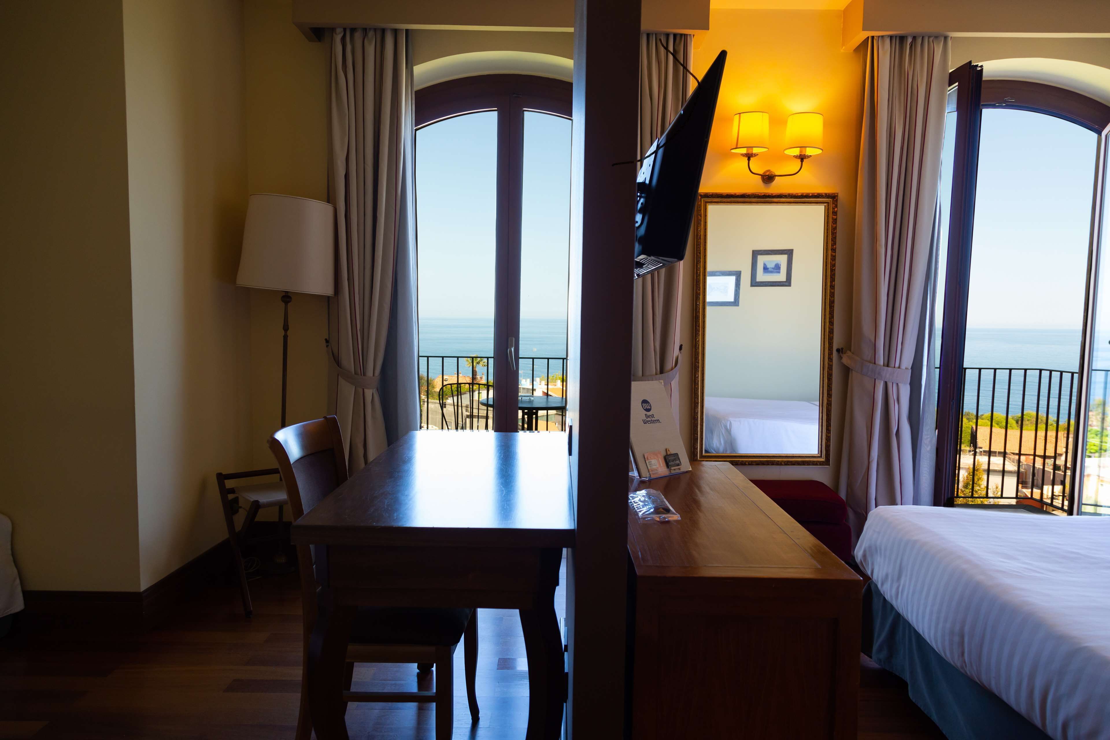 A hotel room with a wooden desk, a bed, a TV, and a balcony with a view of the sea.