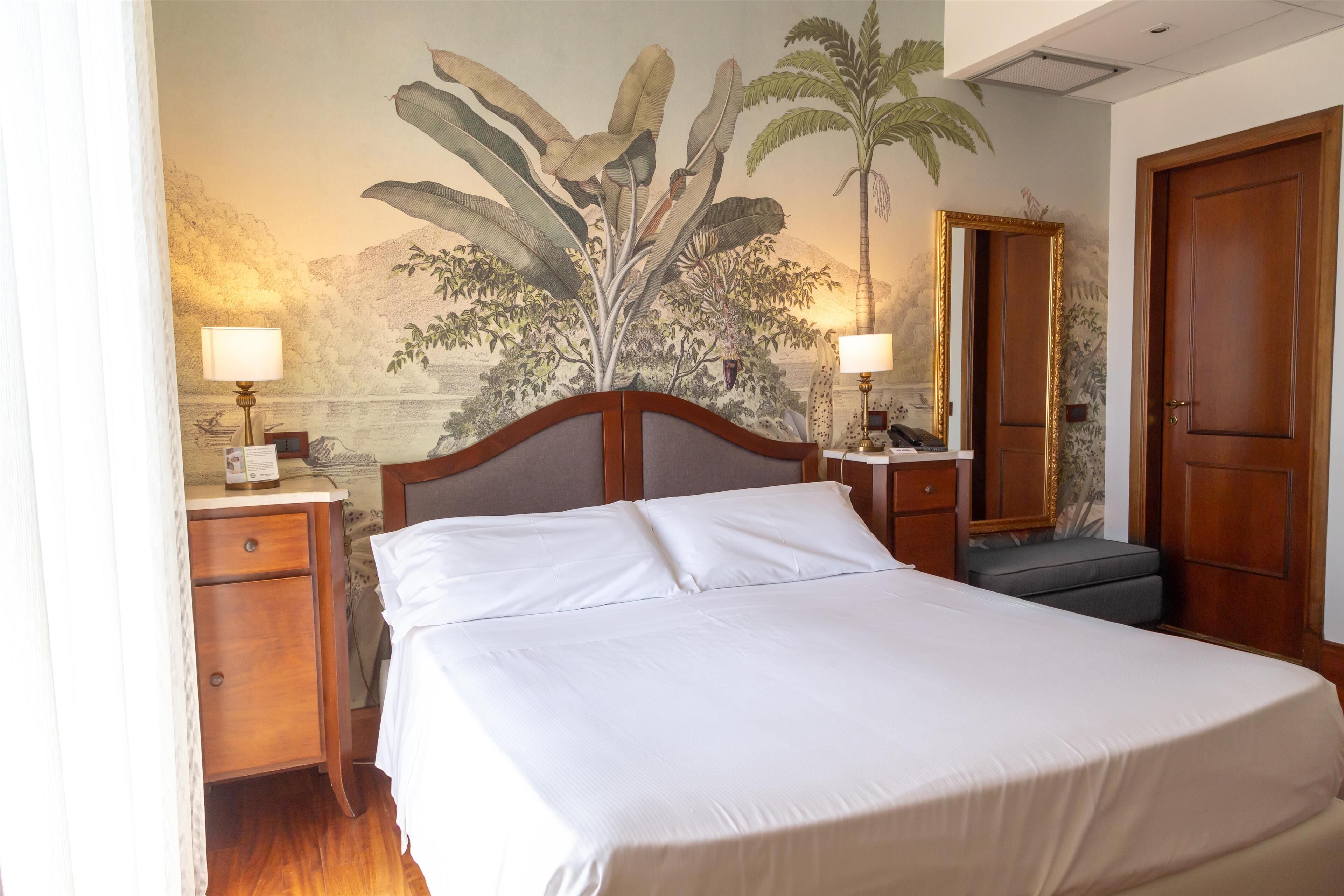A hotel room with a bed, a nightstand, a lamp, a mirror, and a door.
