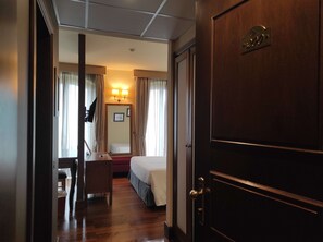 Habitación de hotel con cama, escritorio, silla, espejo y puerta con número de habitación.