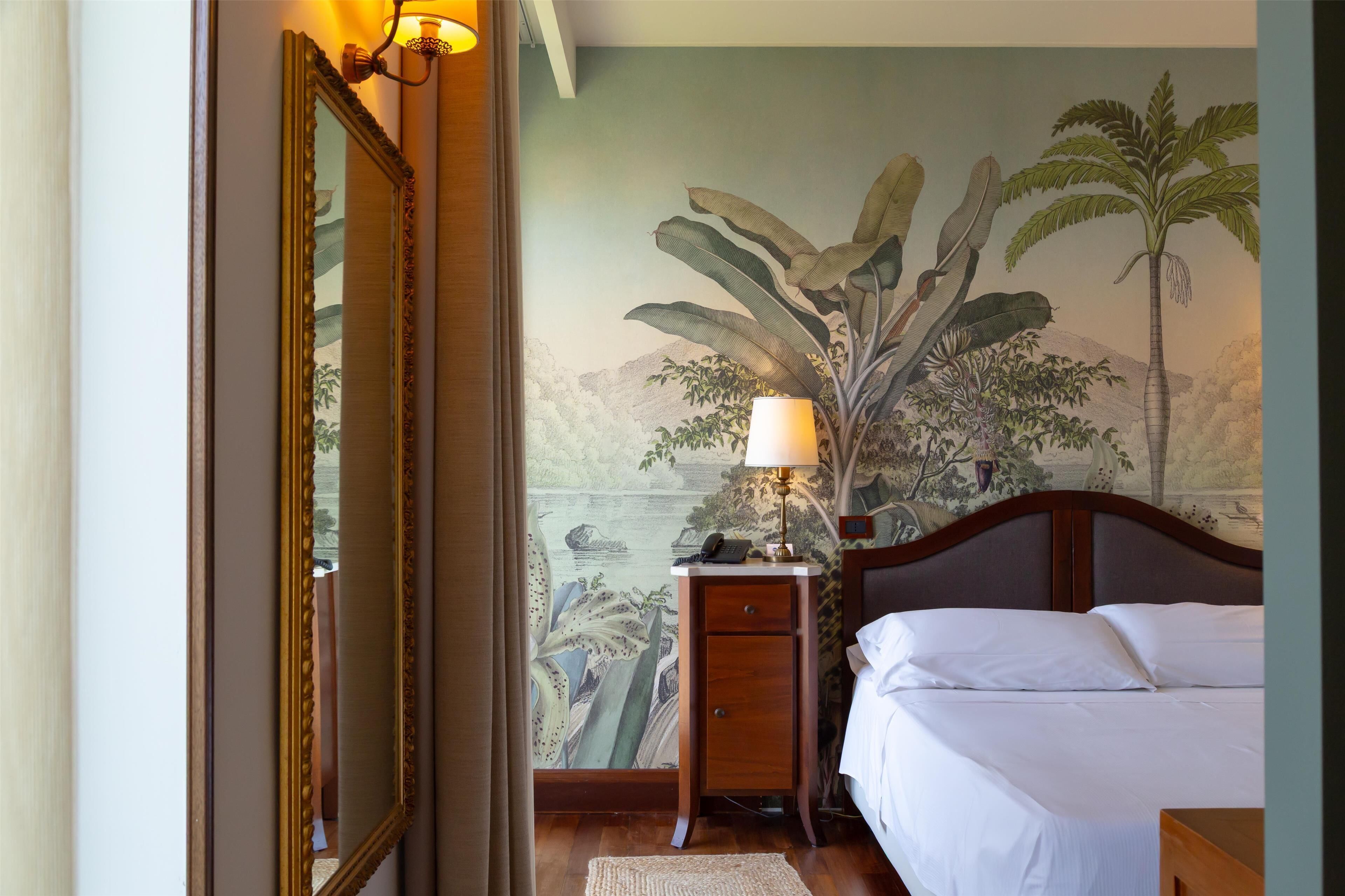 Habitación de hotel con cama, mesita de noche con lámpara, espejo y un mural de paisaje tropical.