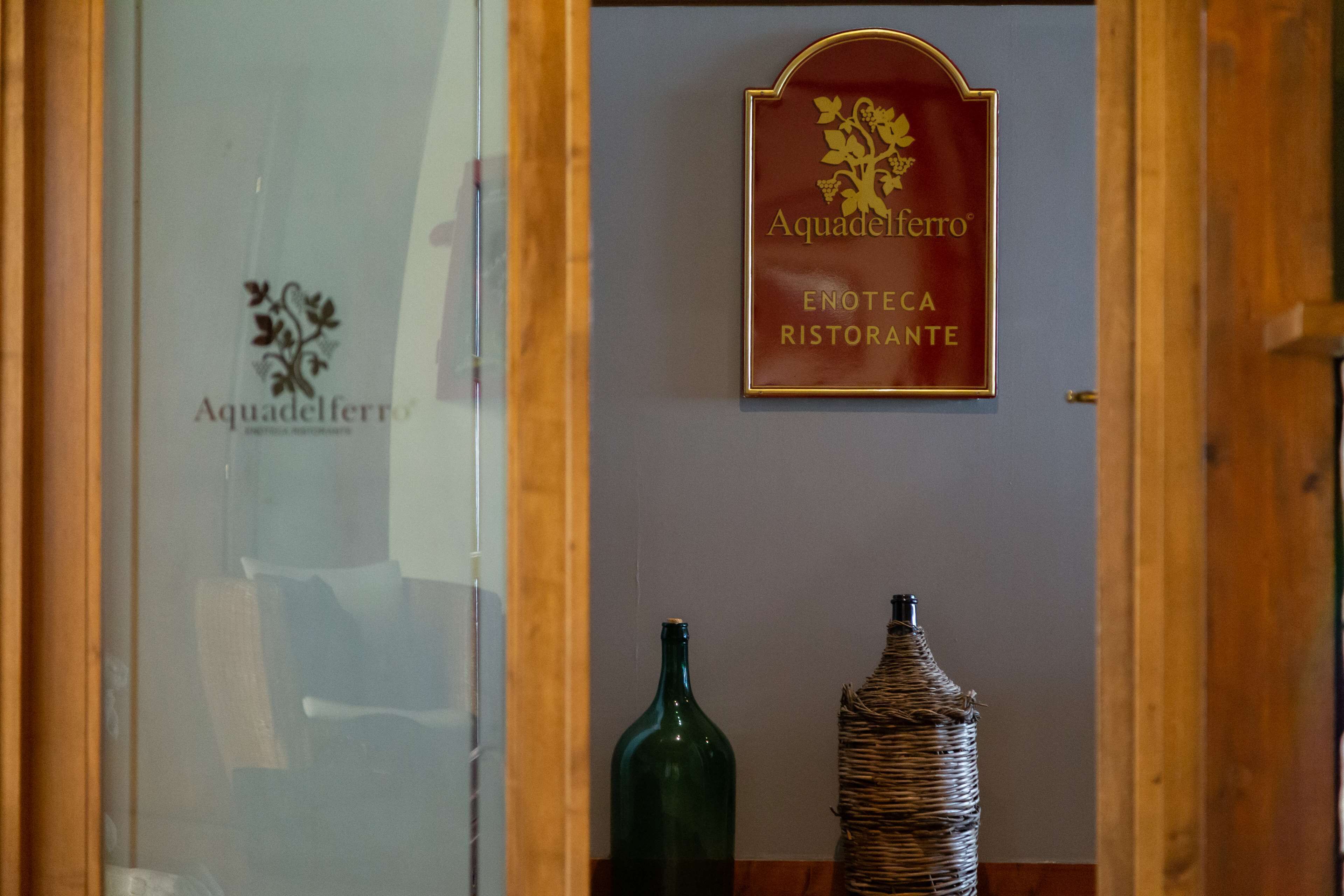 Un letrero de restaurante con el nombre 'Aquaferro' y 'ENOTECA RISTORANTE'. Hay dos botellas, una verde y otra de mimbre, sobre una mesa.