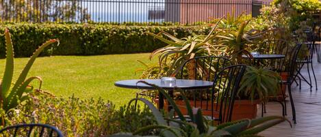 Un patio al aire libre con mesas y sillas, rodeado de plantas en macetas y un césped bien cuidado.