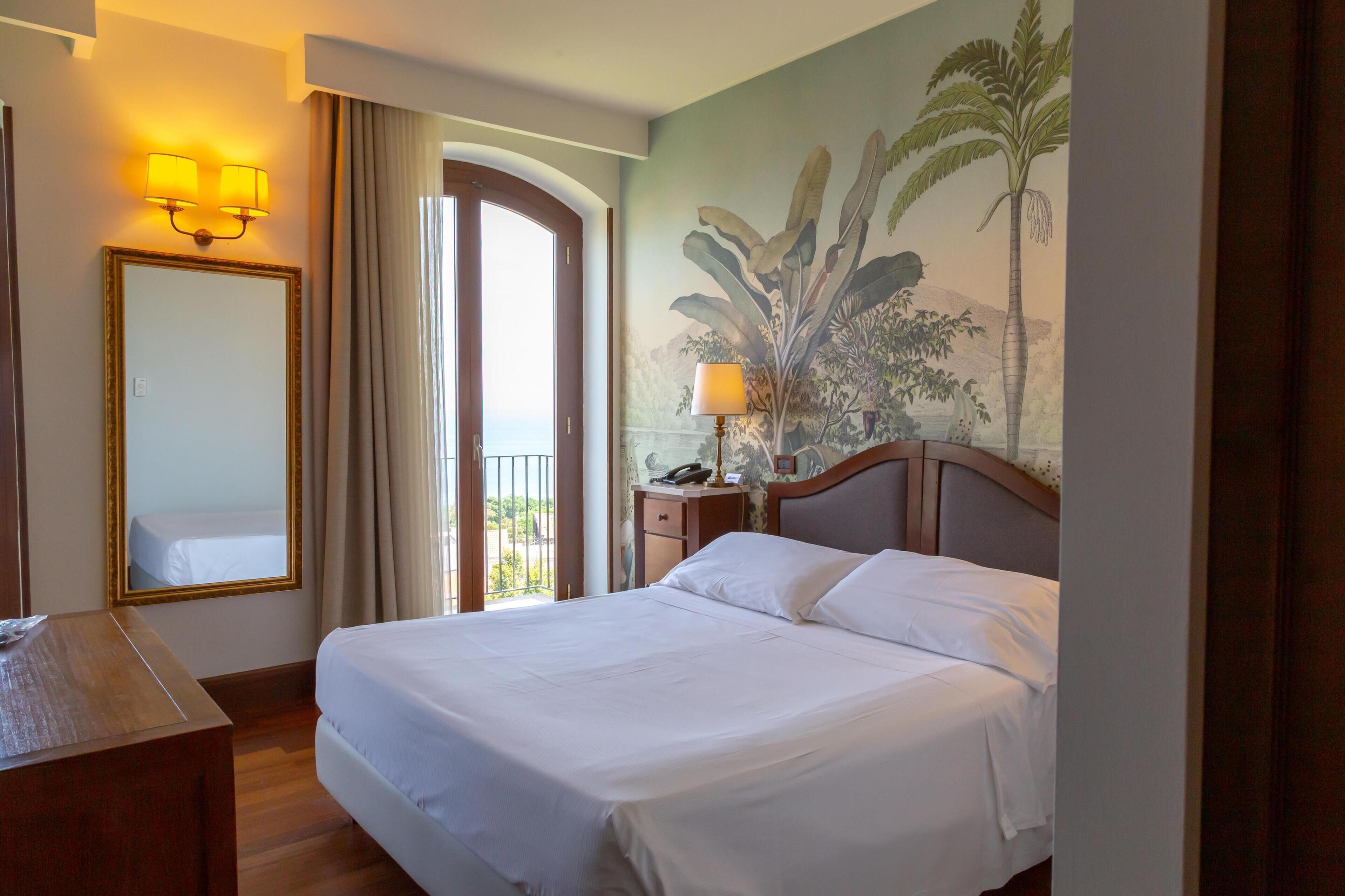 A hotel room with a bed, a desk, a mirror, a nightstand, a lamp, and a large window with curtains.