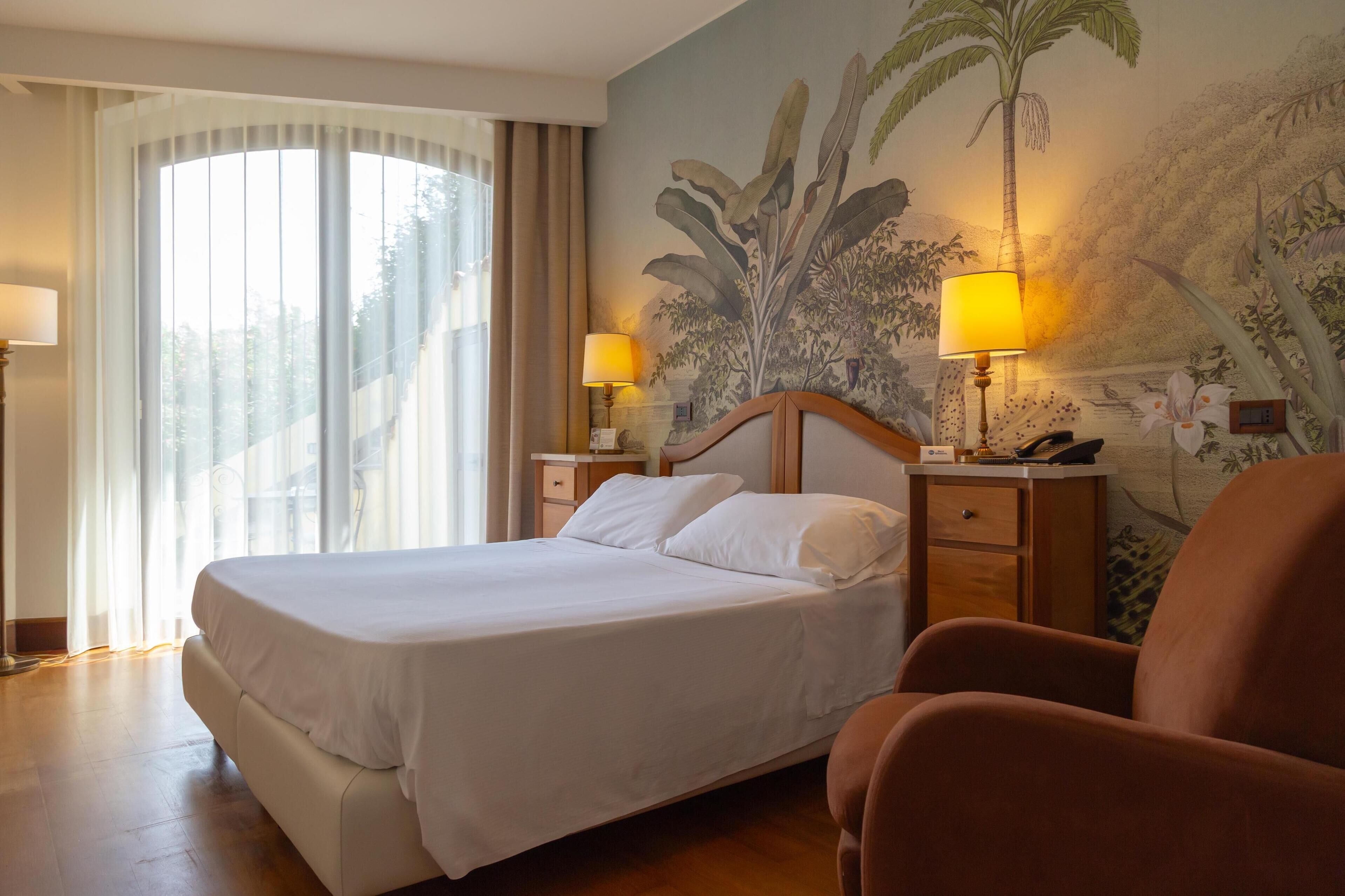 A hotel room with a large bed, two bedside tables with lamps, a chair, and a mural of tropical foliage on the wall.