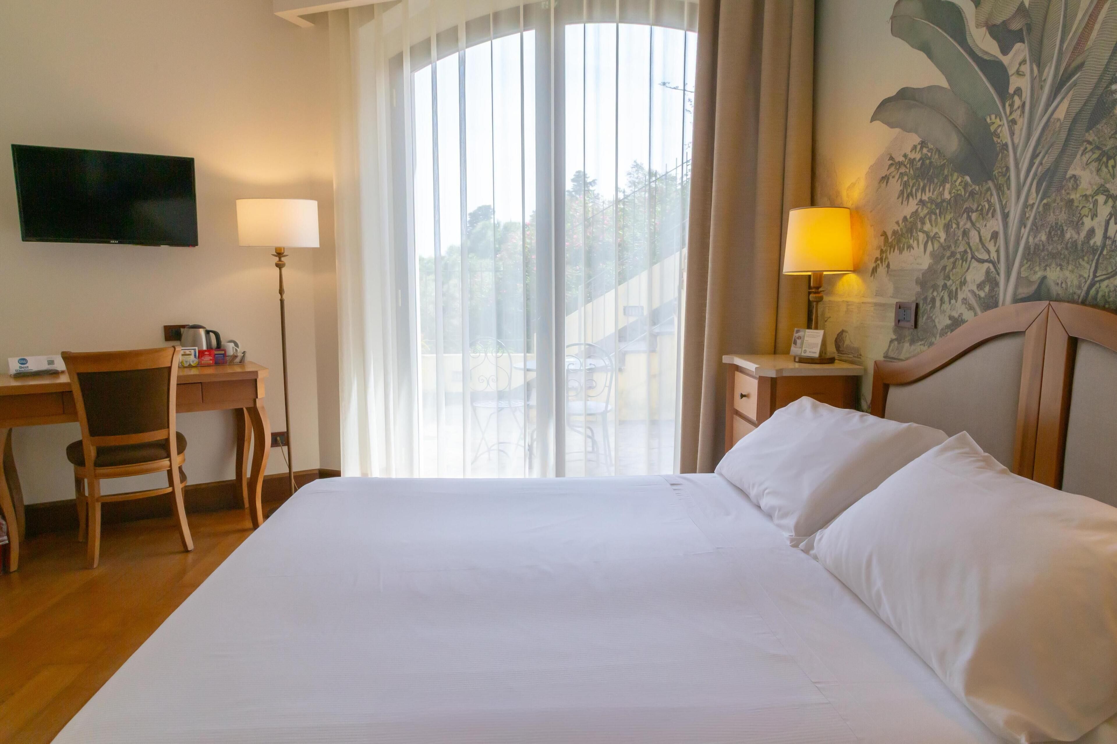 Una camera d'albergo con un letto, una scrivania, una sedia, una televisione e una finestra con tende.
