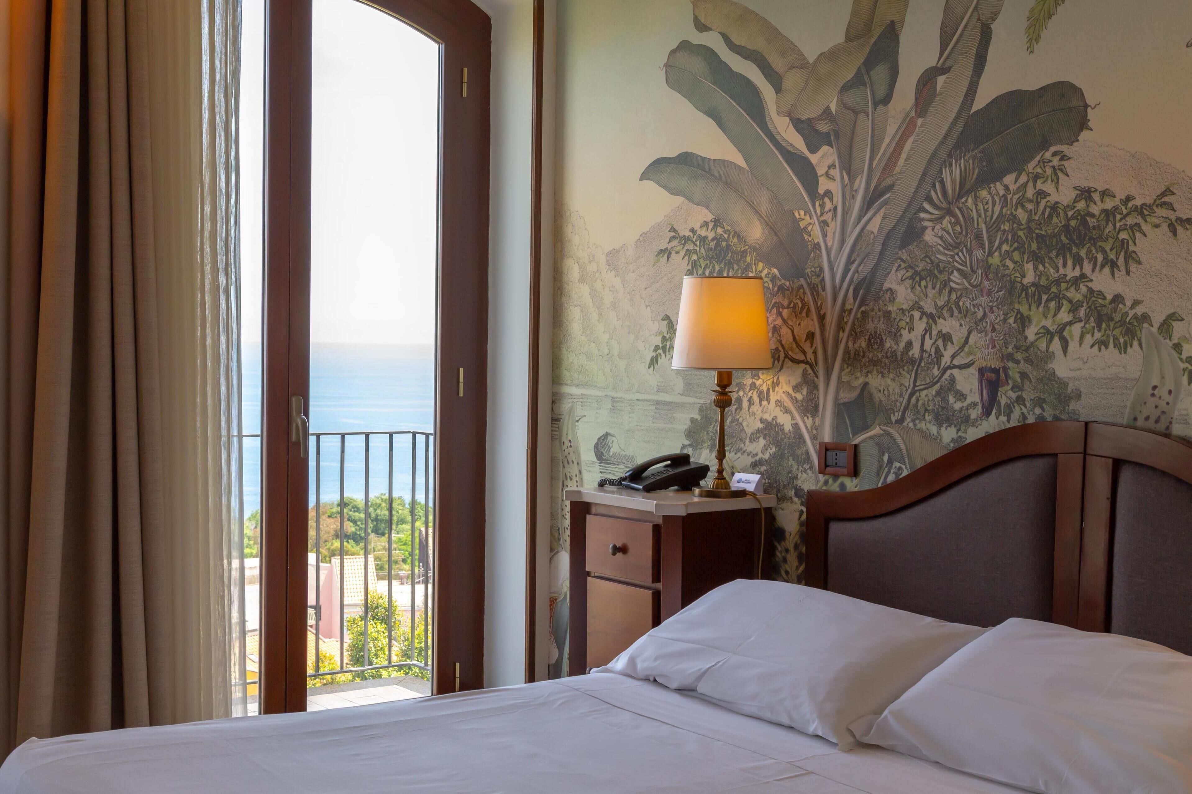 Une chambre d’hôtel avec un lit, une table de chevet avec une lampe, un téléphone et une vue sur l’océan par une grande fenêtre.