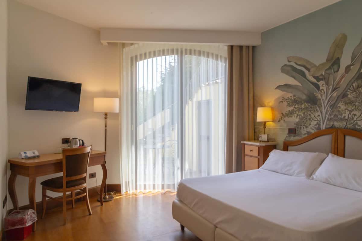 Une chambre d’hôtel comprenant un lit, un bureau avec une chaise, une télévision, une fenêtre avec des rideaux et un tableau au mur.
