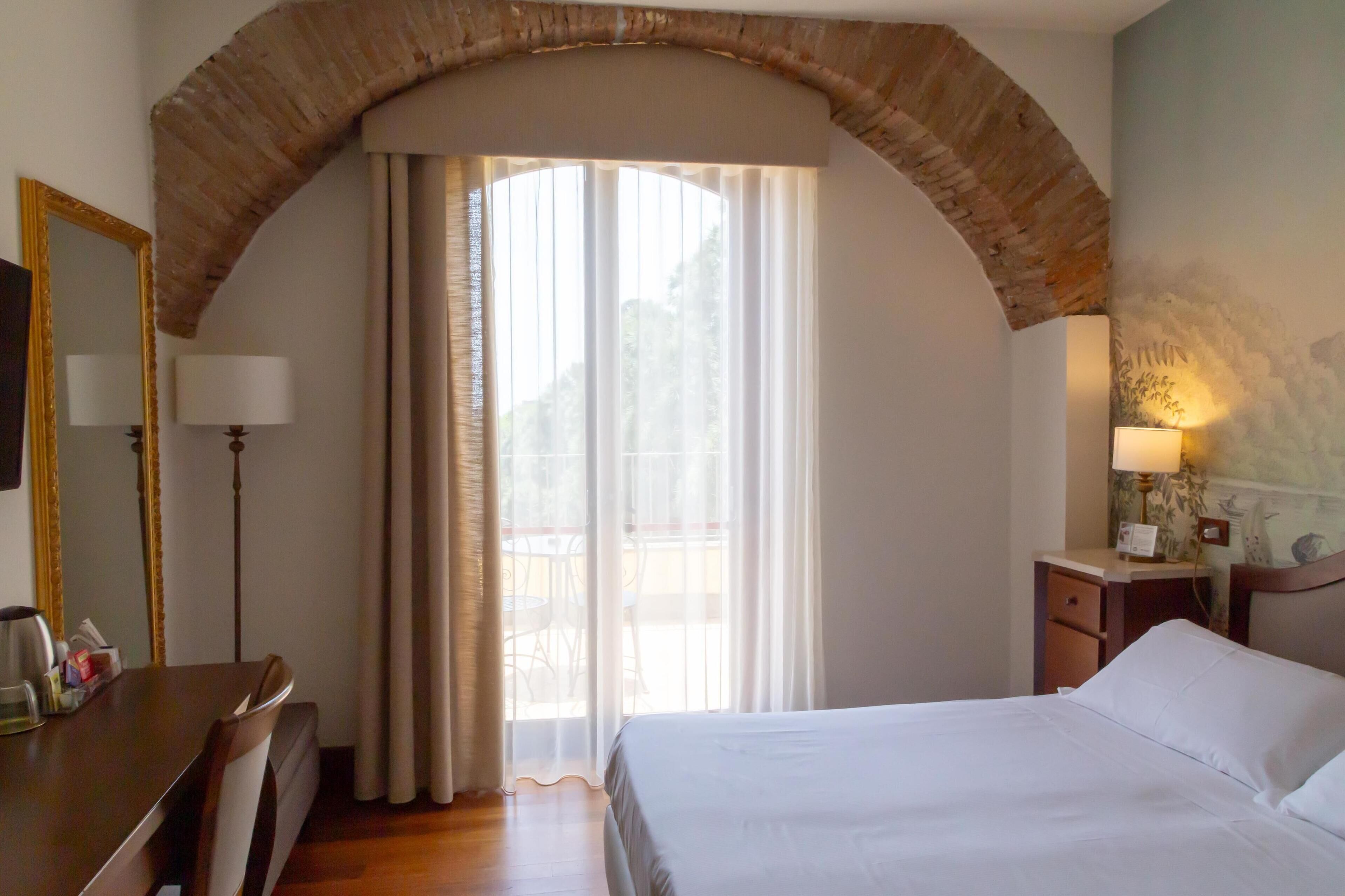 Habitación de hotel con cama, escritorio, silla, lámpara y ventana con cortinas.