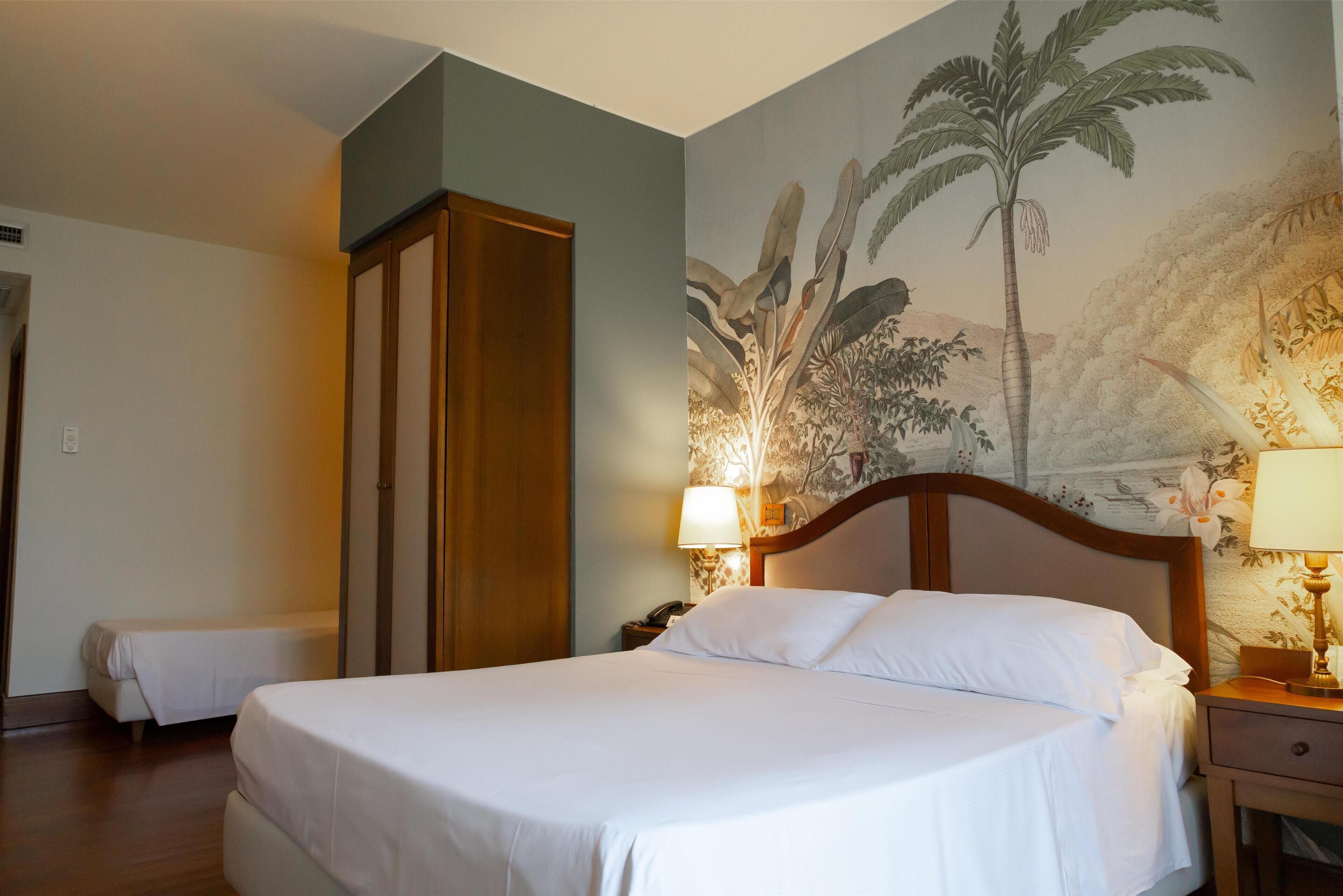 Una habitación de hotel con una cama grande, una mesita de noche con lámpara, un armario y un mural con follaje tropical en la pared.
