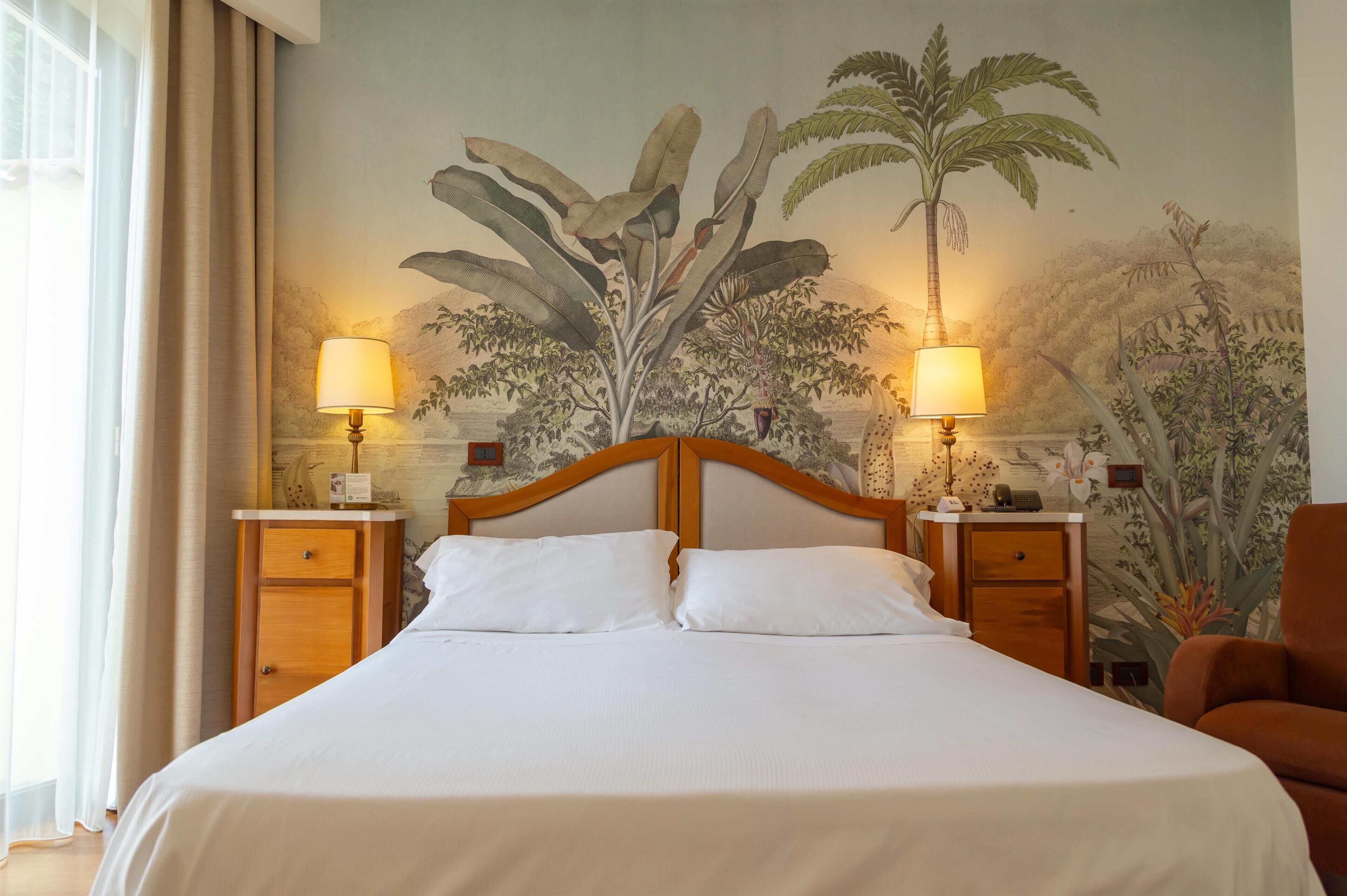 A hotel room with a large bed, two bedside tables with lamps, a wooden headboard, and a mural of tropical plants on the wall.