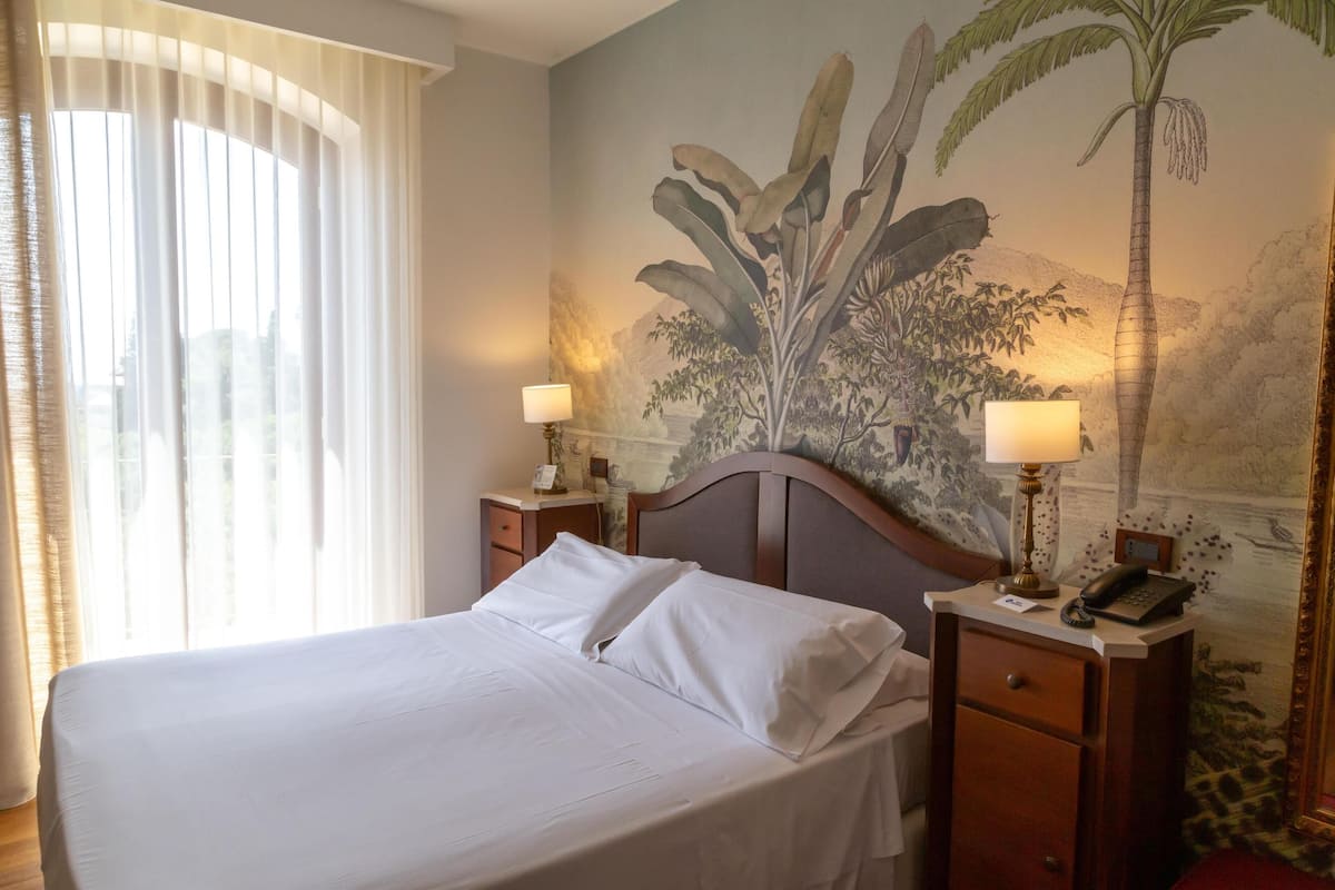 Une chambre d’hôtel avec un lit, deux tables de chevet avec des lampes, une petite table et une fresque représentant des plantes tropicales sur le mur.