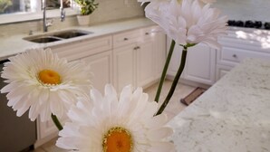 A kitchen with white cabinets, a sink, and a window. There are three white flowers in a vase on the counter. - Star of the Sea Guest House (White Rock)