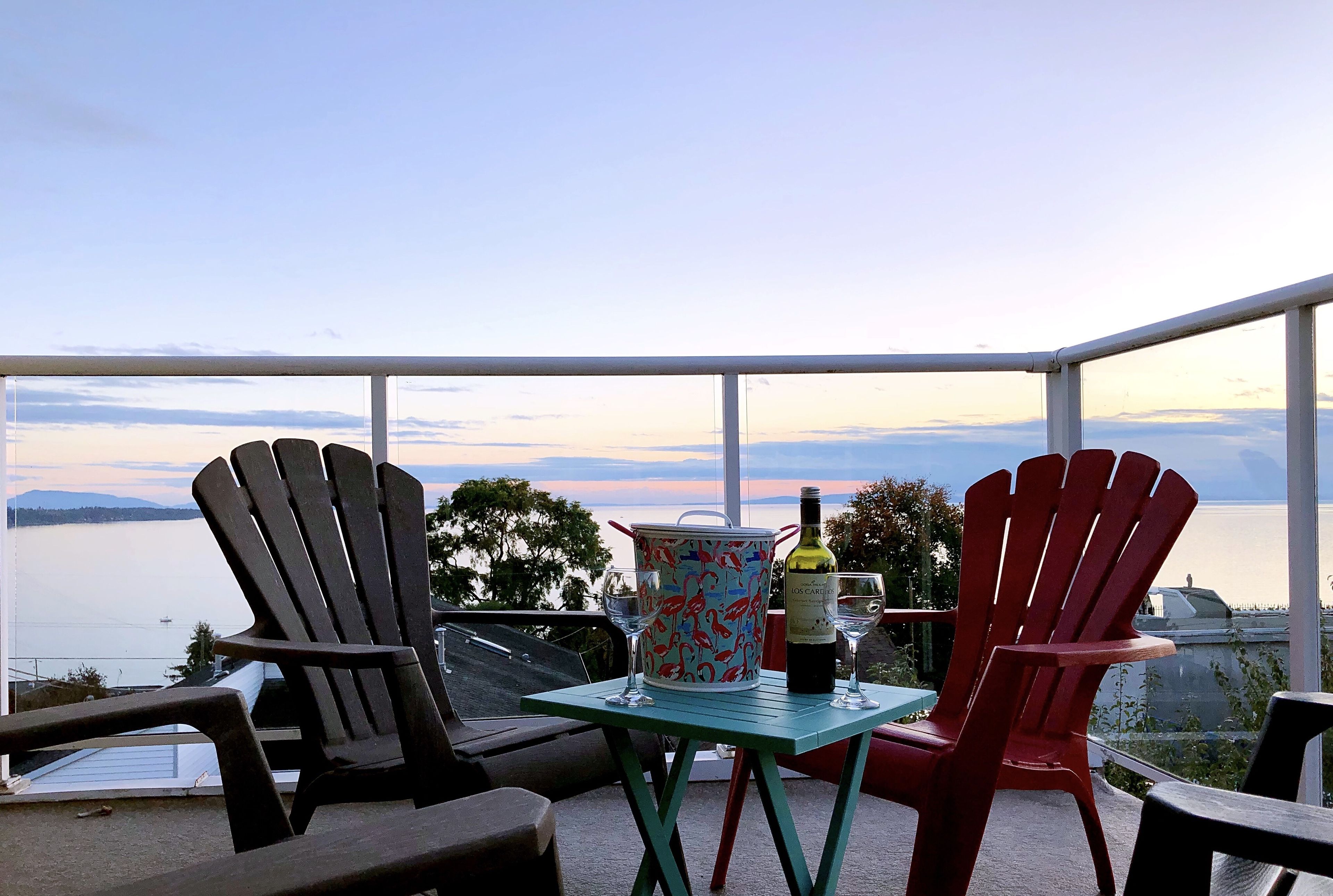 A balcony with two Adirondack chairs, a table with a wine bottle and glasses, and a view of the water.