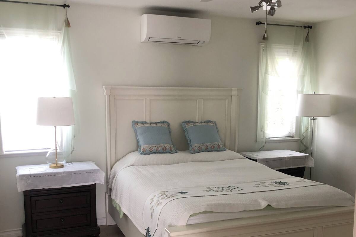 A bedroom with a white bed, two bedside tables, a ceiling fan, and an air conditioning unit.