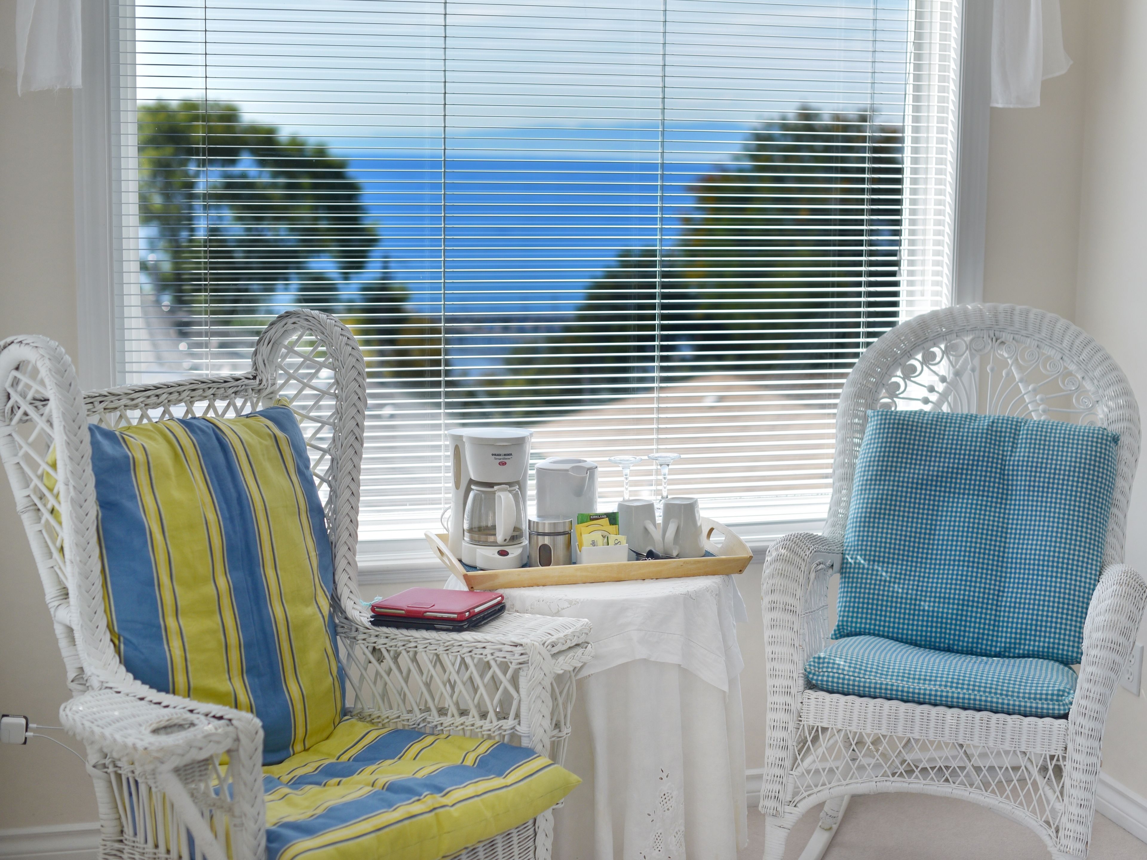 Quarto com duas cadeiras de vime, uma com almofada listrada e outra com almofada xadrez, uma mesa pequena com uma cafeteira e uma bandeja de objetos, e vista para o mar através de grandes janelas com persianas.