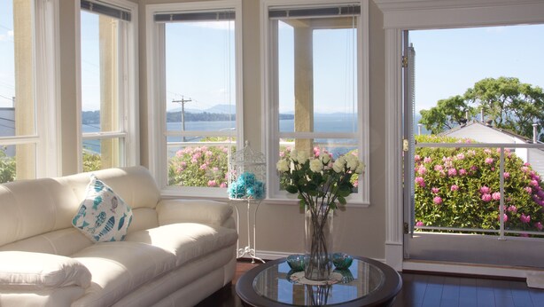 A room with a white sofa, a glass table, and a view of a garden with pink flowers through a large window. - Star of the Sea Guest House (White Rock)