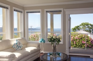 Quarto com sofá branco, mesa de vidro e vista para um jardim com flores rosa através de uma grande janela.