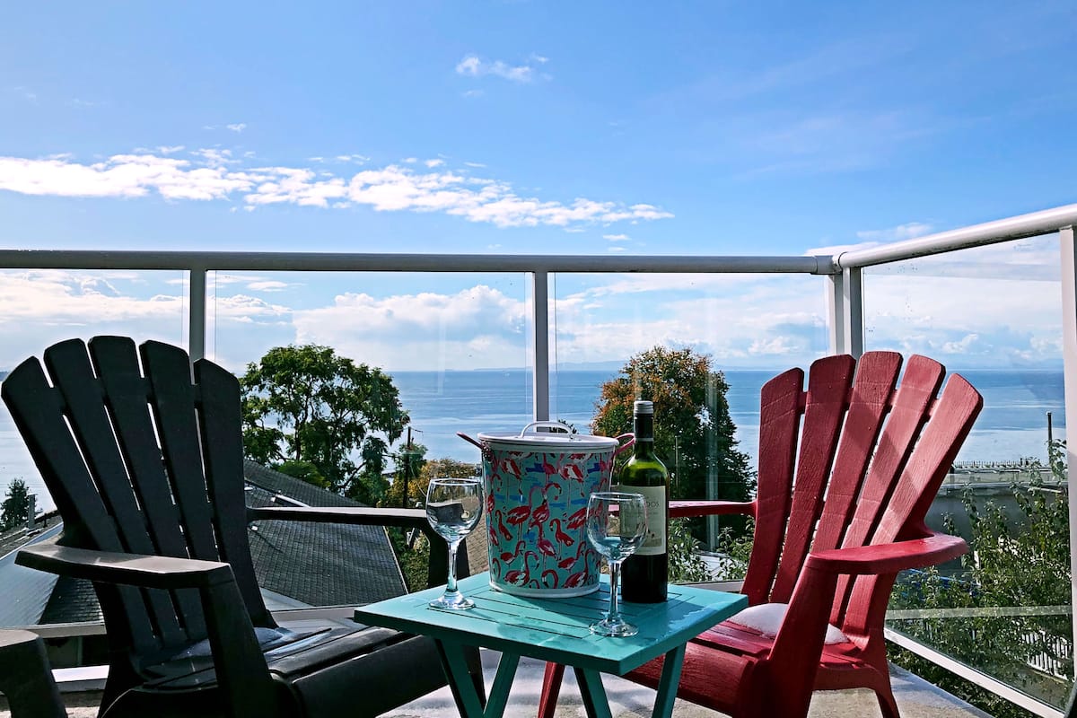 A balcony with two Adirondack chairs, a small table with a decorative vase, a wine bottle, and a glass. The view includes a body of water and distant land.