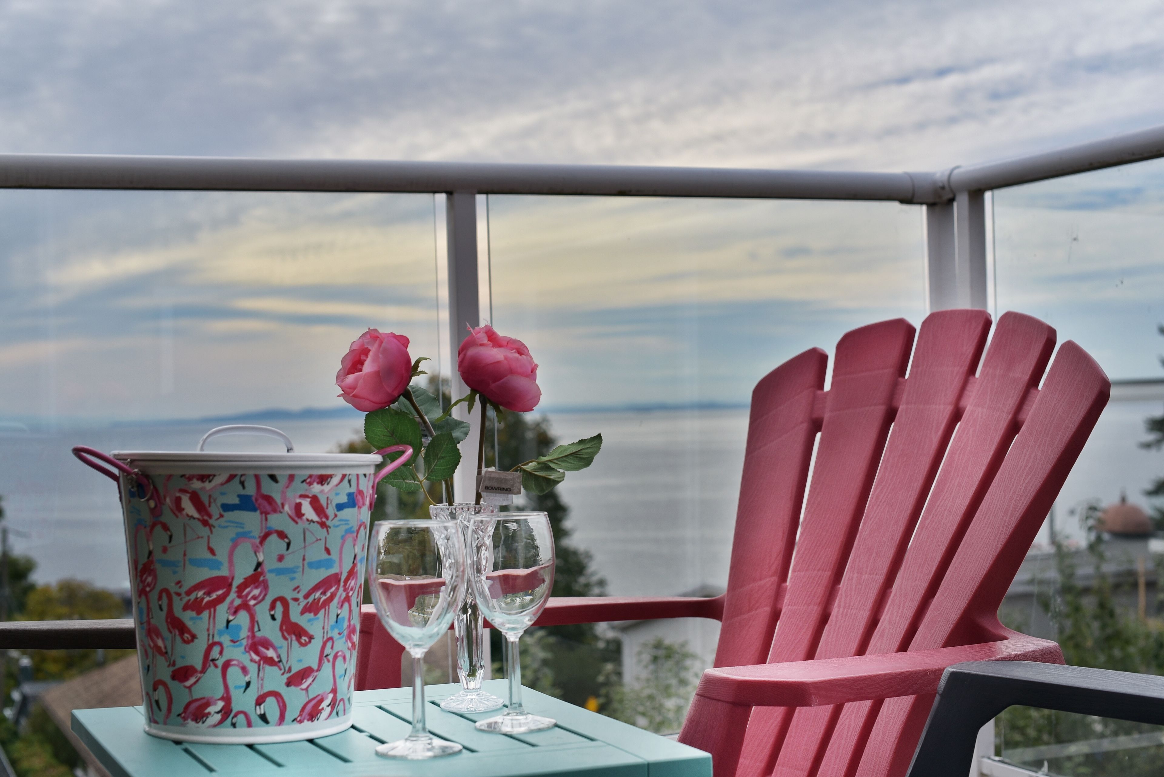 A balcony with a view of the sea, a pink chair, a turquoise table, a bucket with flamingos, and two wine glasses.