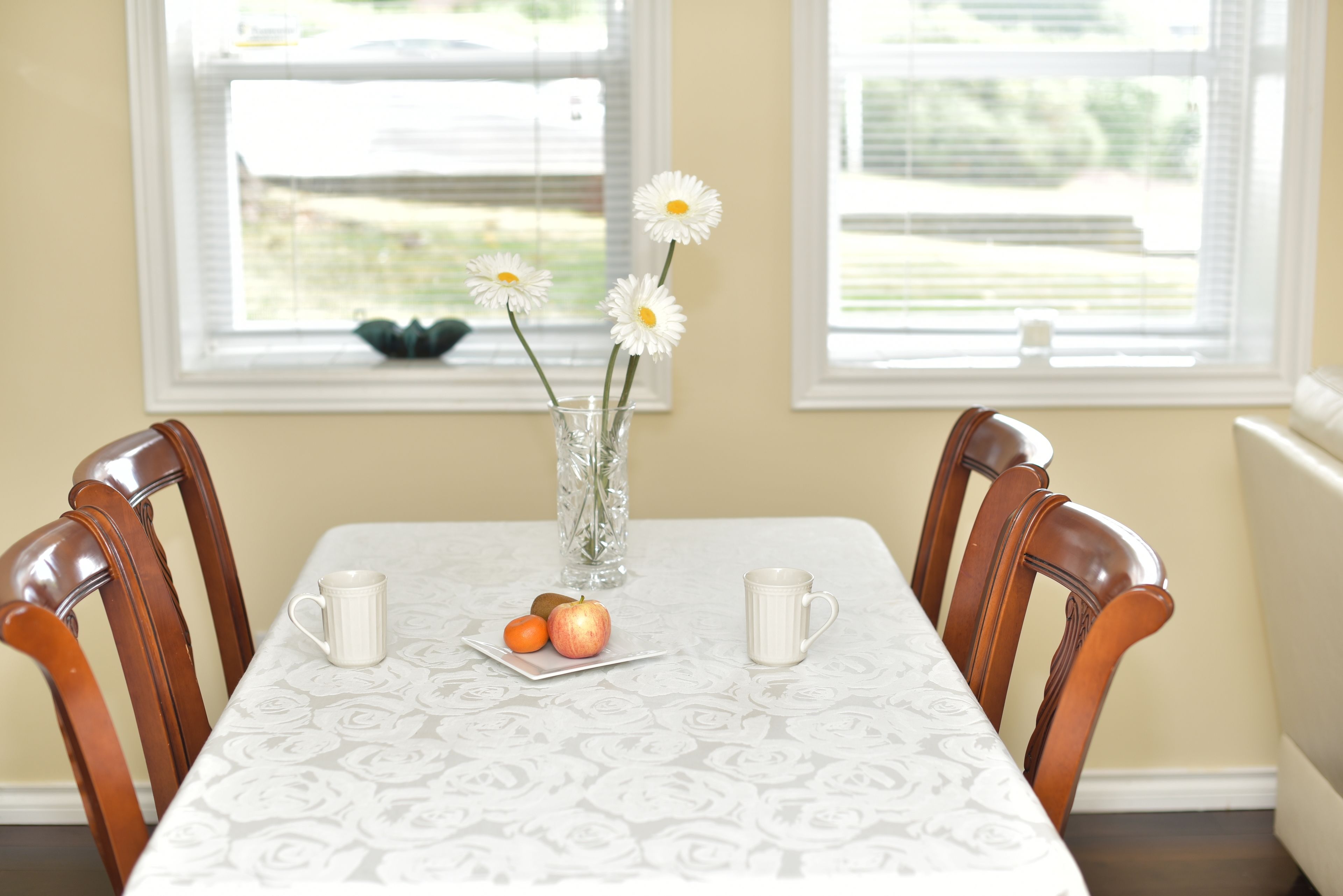 Una mesa de comedor con mantel floral, dos tazas blancas, un jarrón con flores blancas y un plato con una manzana y un tomate.