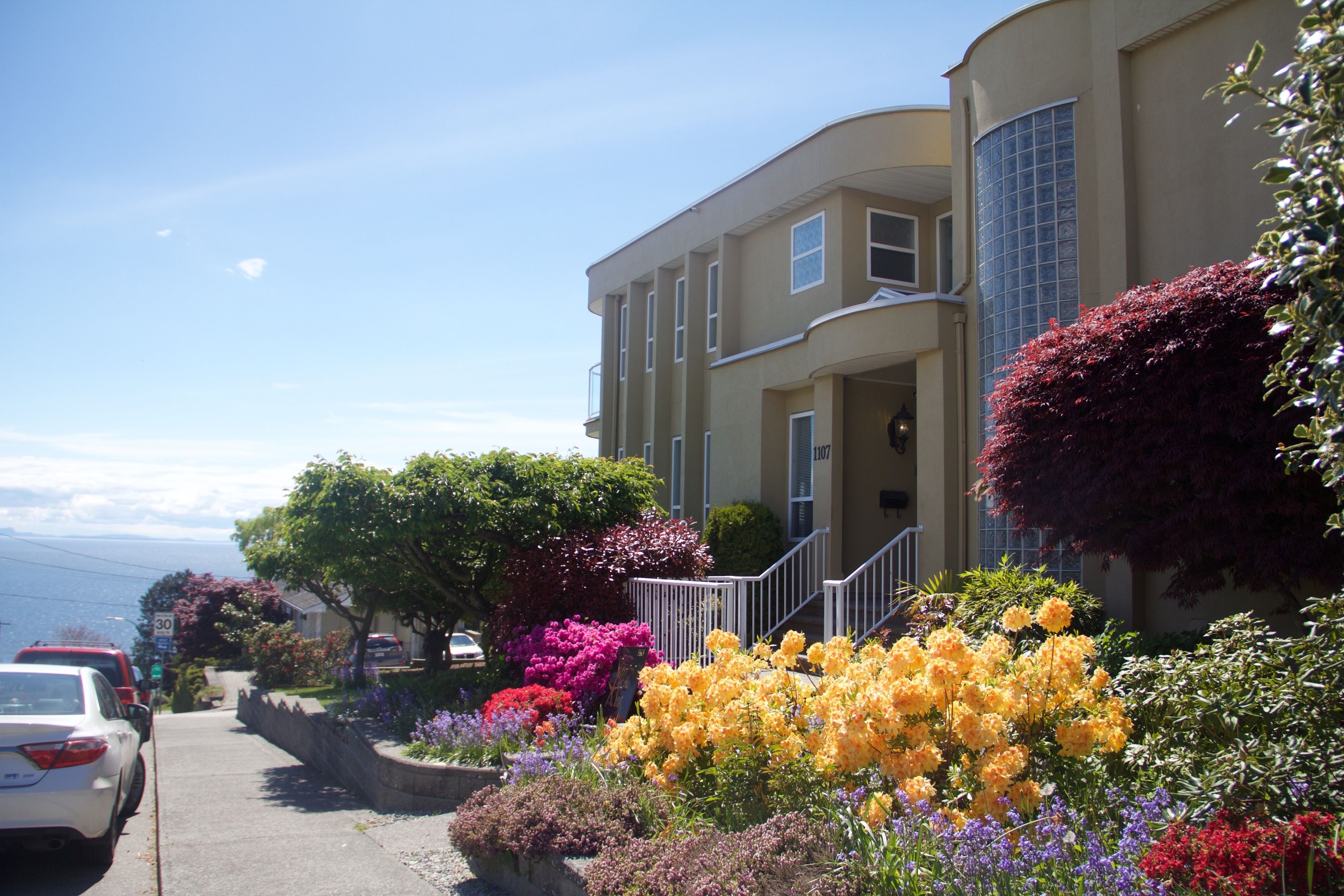 A residential building with a curved facade, a visible staircase, and a variety of colorful flowering plants in the front yard.