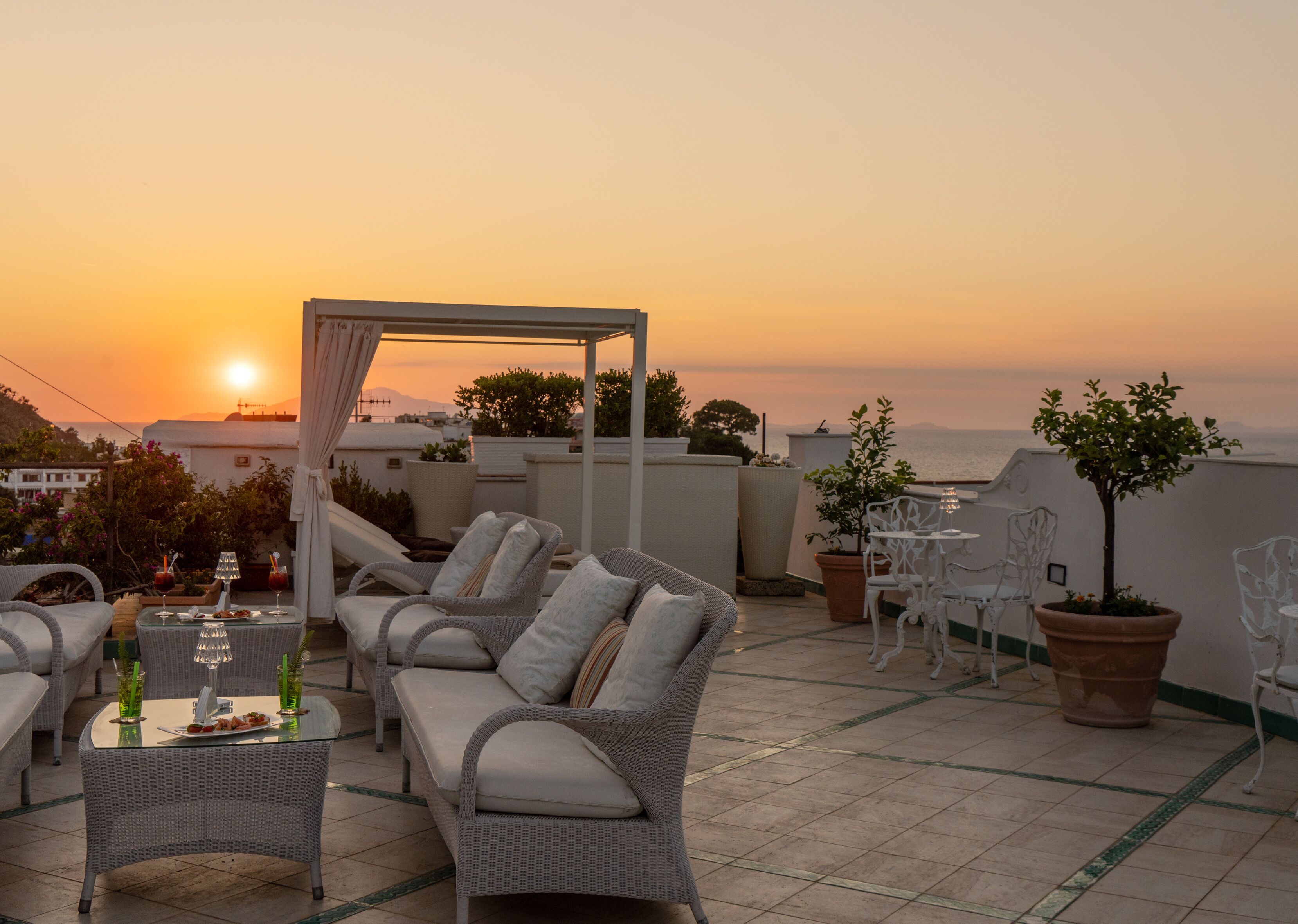 A rooftop terrace with wicker furniture, a table with drinks, and potted plants.